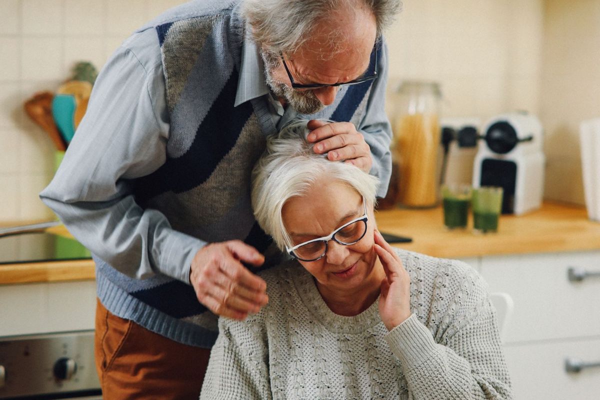 Een ouder echtpaar in hun keuken; hij staat achter haar met een hand op haar schouder terwijl zij nadenkend voor zich uit kijkt